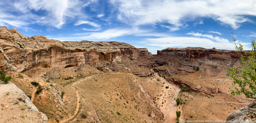 A small hiker stands on a steep canyon trail above a deep basin of layered cliffs and eroded slopes.