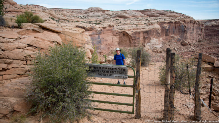 A hiker stands beside a green gate and rock-art protection sign in a sandstone canyon.
