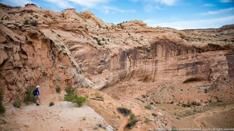 A hiker follows a sandy path beneath a high, layered sandstone cliff.