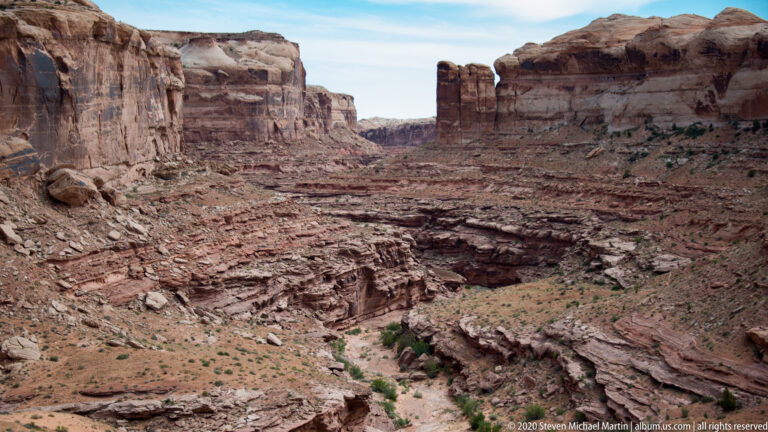 A deep sandstone canyon extends between layered cliffs above a rocky, sandy wash.