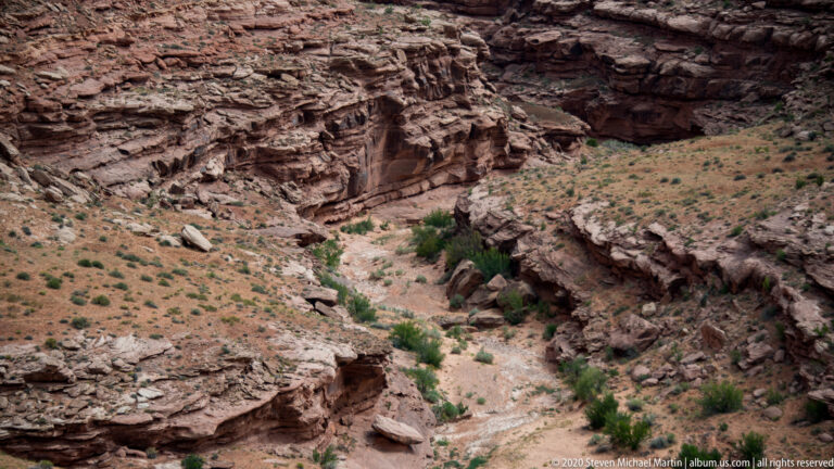 A pale wash winds between broken red sandstone shelves and scattered shrubs.