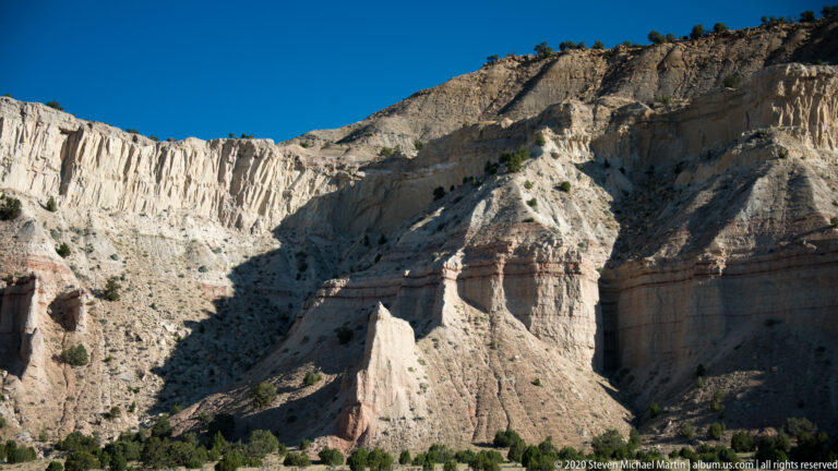 Sunlit pale cliffs show deep vertical gullies, angular fins, and a darker caprock.