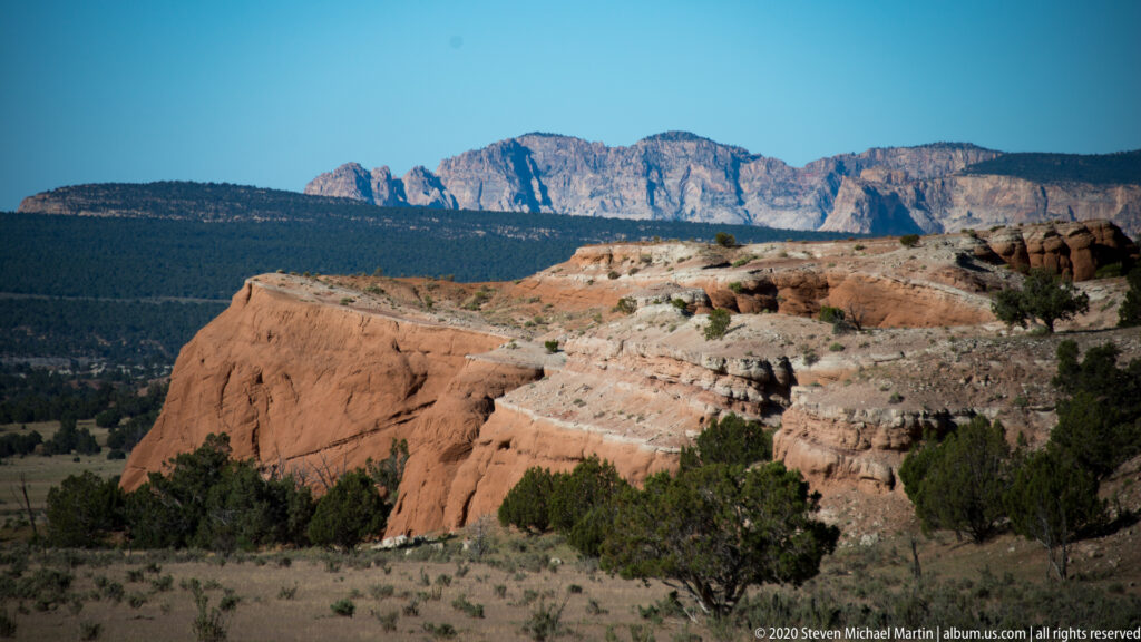 Red sandstone slopes toward junipers, with pale cliffs and blue mountains behind.