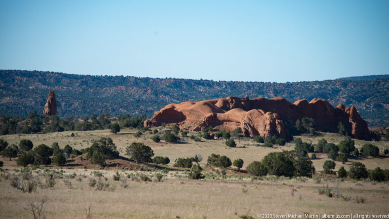 A narrow red spire stands left of a low rock ridge beyond a dry meadow.