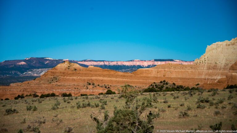 Red and cream rock bands cross the basin below a pale cliff and blue sky.