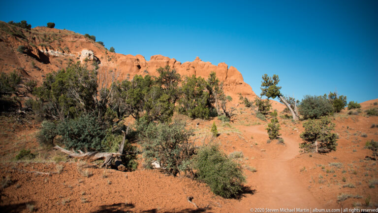 A sandy path winds beneath junipers toward orange sandstone formations on a dry slope.