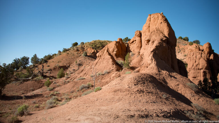 A narrow orange sandstone pillar rises from a red slope before pale cliffs.