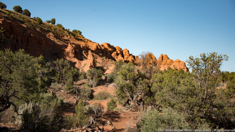 A low orange sandstone ridge crosses a juniper-covered slope beneath a clear sky.