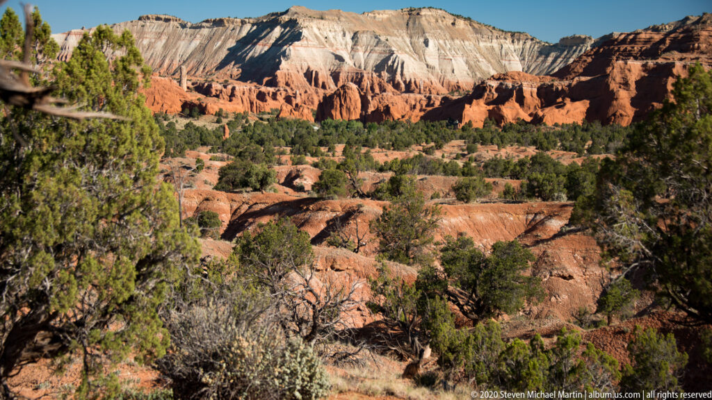 Red gullies lead toward a long pale cliff band above scattered junipers and shrubs.
