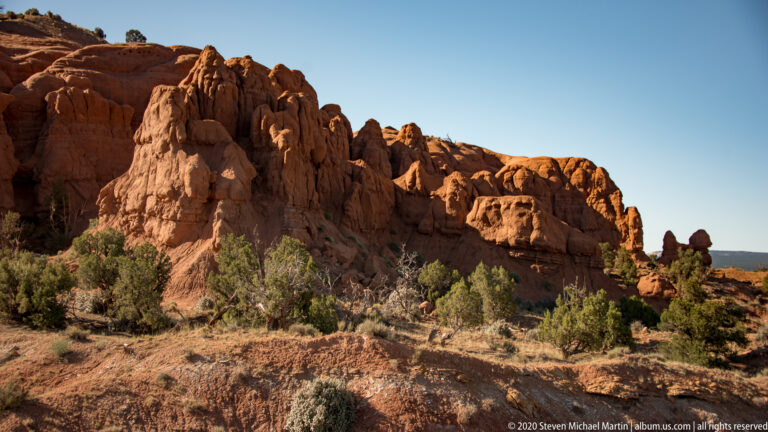 A rugged red sandstone wall rises beside a sandy path and low desert shrubs.