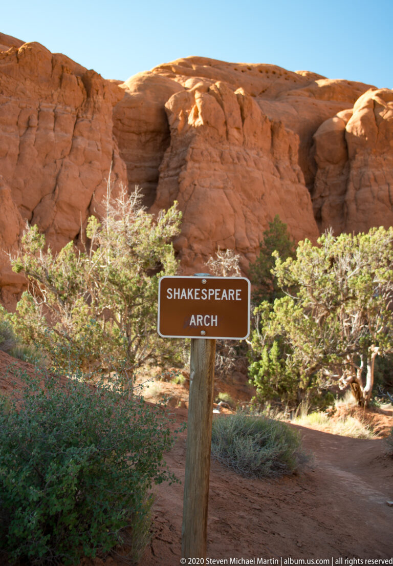 A brown sign marked “Shakespeare Arch” stands before a high red sandstone wall.
