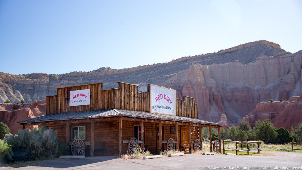 A timber storefront with “RED DIRT” signs stands before tall pink sandstone cliffs.