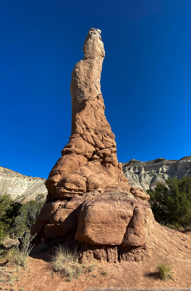 A tall layered sandstone spire rises above a red base with pale cliffs behind it.