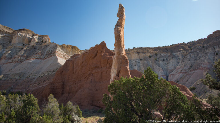 A narrow red sandstone pinnacle rises before pale grooved cliffs and junipers.