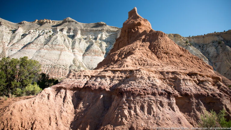 A fluted red sandstone mound rises before pale layered cliffs under a clear blue sky.