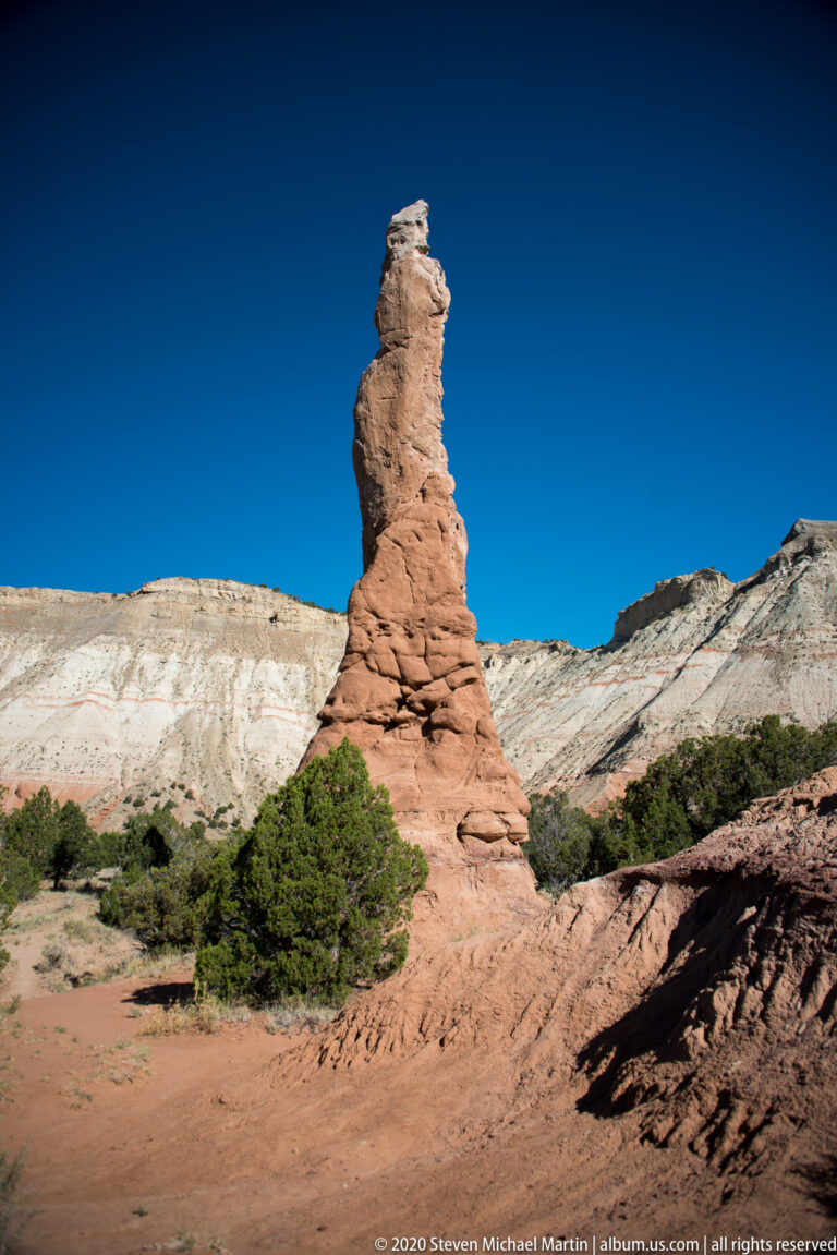 A solitary red sandstone spire rises from a broad eroded base before pale cliffs.