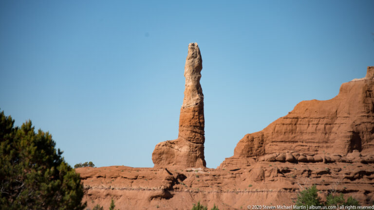 A narrow sandstone chimney rises from a broad red ridge beside a green tree.