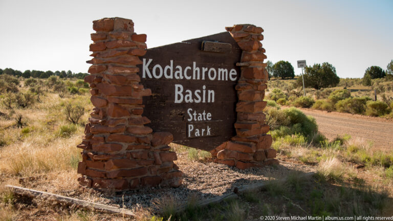 A dark sign reading “Kodachrome Basin State Park” stands between stone pillars beside a dirt road.
