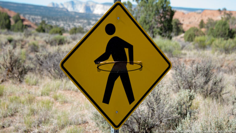 A yellow diamond sign with a black walking figure stands before blurred shrubs and red rock.