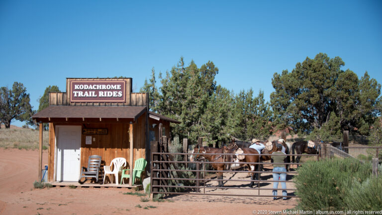 A Kodachrome Trail Rides shelter stands beside saddled horses in a fenced pen, with a person near the gate.