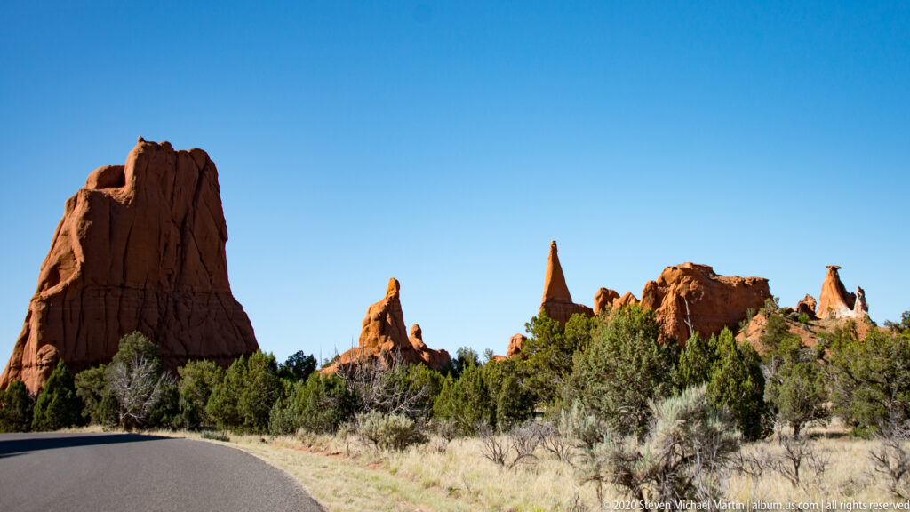 A road curves past a large red sandstone tower and smaller pinnacles above juniper scrub.