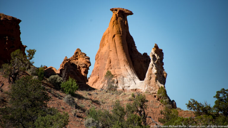 A capped sandstone spire rises above brush and smaller red rock formations on a slope.