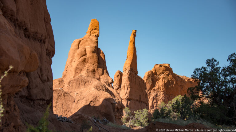 Multiple red sandstone towers and a thin spire rise beyond a shaded rock wall.