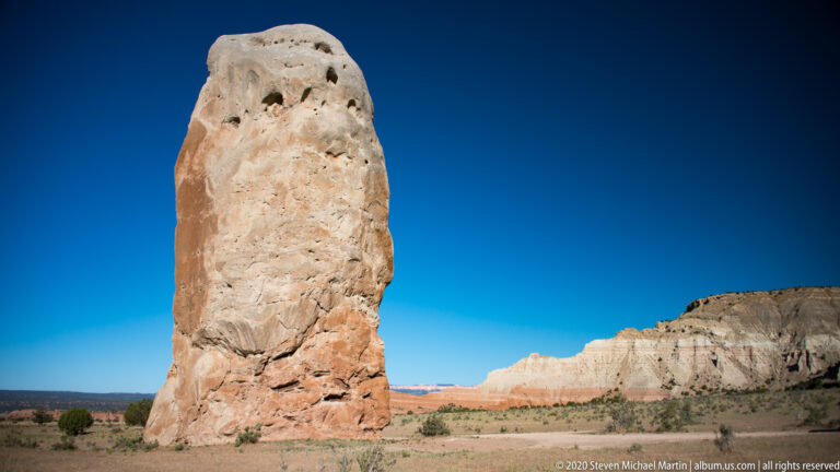 A pale pitted sandstone pillar rises alone on a flat plain before distant layered cliffs.