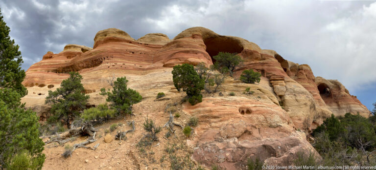 A banded sandstone wall with dark alcoves rises above junipers and dry brush beneath clouds.