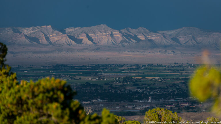 A developed valley lies beneath a long pale ridge at dusk, viewed past softly blurred shrubs on a hillside.