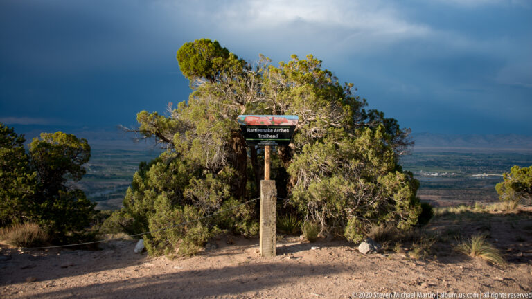 A Rattlesnake Arches Trailhead sign stands among junipers above a broad valley beneath a dark, cloud-filled sky.