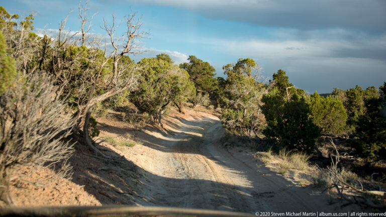 A narrow dirt road curves through juniper and scrub beneath a cloudy sky.