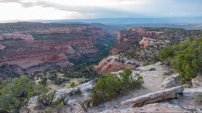 A rocky overlook faces a deep red sandstone canyon with a broad promontory, layered cliffs, and scrub-covered slopes.