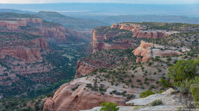 A large sandstone promontory projects above a winding canyon, with layered red cliffs and a distant blue plateau.