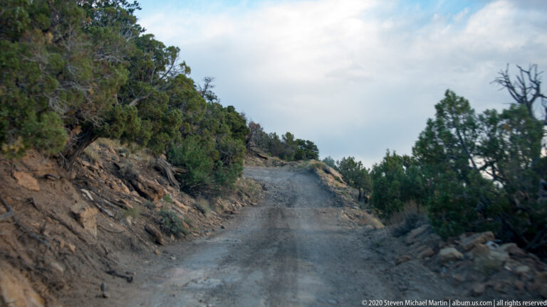 A rough gravel road bends along a rocky hillside between shrubs and small trees under an overcast sky.
