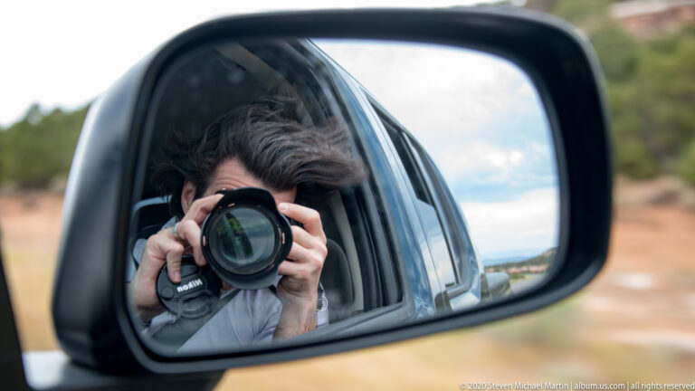 A camera-holding photographer appears in a vehicle side mirror, framed by its black edge.