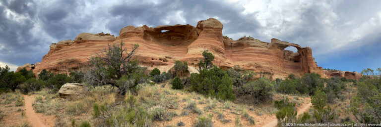 A curved banded sandstone ridge with small cavities rises above juniper scrub and a foreground boulder.