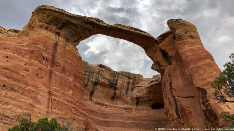 A broad sandstone arch bridges two orange rock walls beneath a cloudy sky.