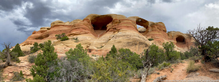 A large dark alcove and smaller openings interrupt a rounded, striped sandstone ridge.