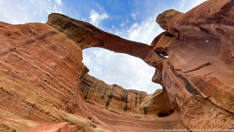 A sandstone arch frames a recessed wall and dark cavity beneath a cloudy sky.