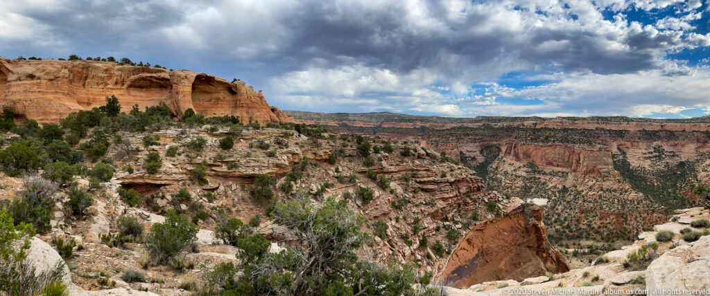 A natural rock span sits above a deep green ravine beneath layered red cliffs.