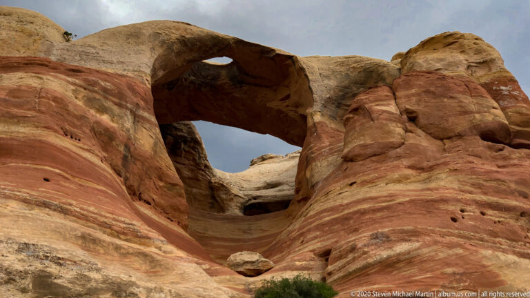 A thick sandstone arch frames pale rock and cloudy sky through a rounded opening.