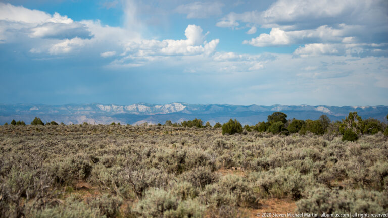 Sagebrush and junipers cover a plateau below distant snow-covered mountains and cumulus clouds.