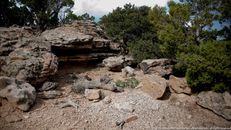 A low sandstone ledge shelters a dark shallow recess amid gravel and juniper shrubs.