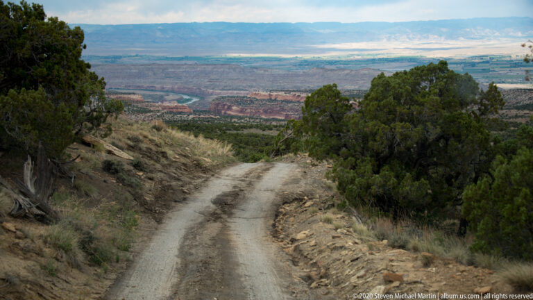 A rutted dirt road descends between junipers toward distant mesas and an open valley.
