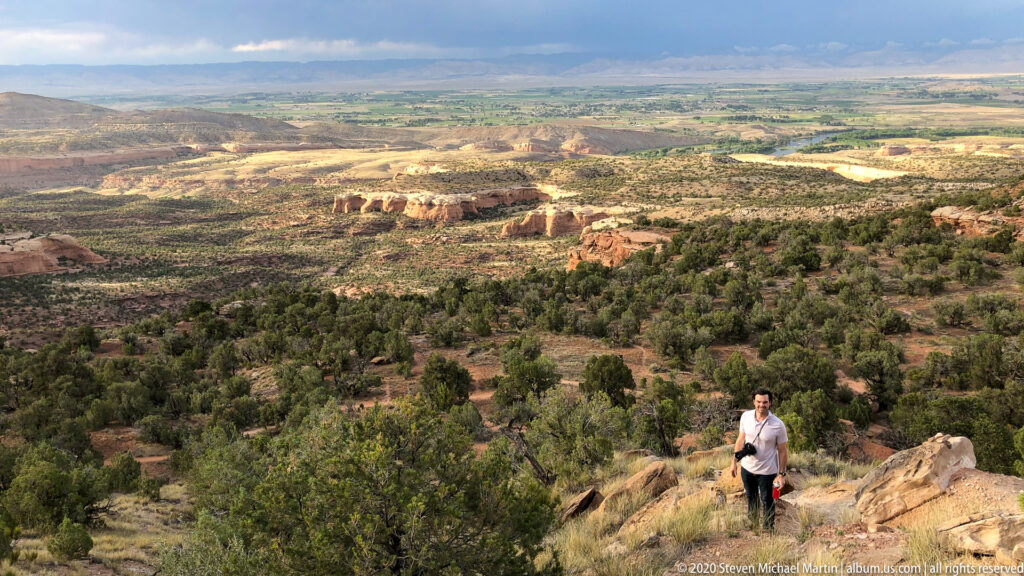 A visitor stands on foreground rocks above a broad scrub-covered valley and red mesas.