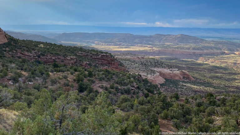 A red cliff band overlooks patchy shrubland, distant mesas, and hazy ridges.