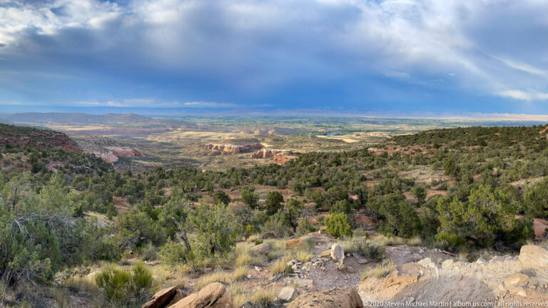 A curtain of rain falls beneath dark clouds over distant mesas beyond dry foreground scrub.