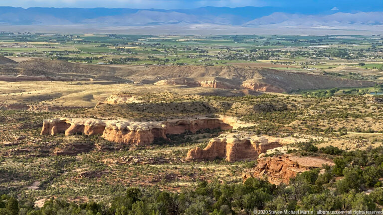 Red sandstone ledges rise from scrubland above a broad cultivated valley and distant mountains.