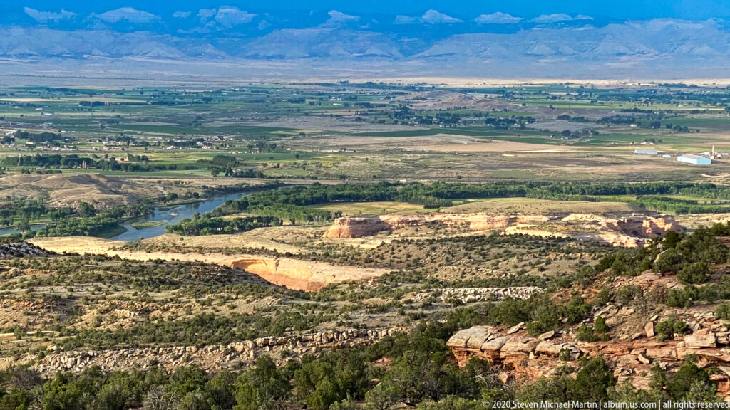 A river corridor winds through a green valley below red canyon benches and distant mountains.